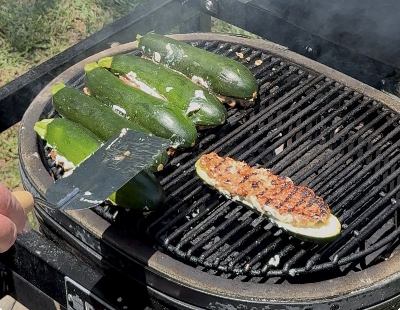 Several zucchinis stuffed with a white cheese mixture are being grilled on a barbecue. One zucchini half is flipped, showing grill marks. A spatula is lifting another zucchini slice. Smoke rises from the grill.
