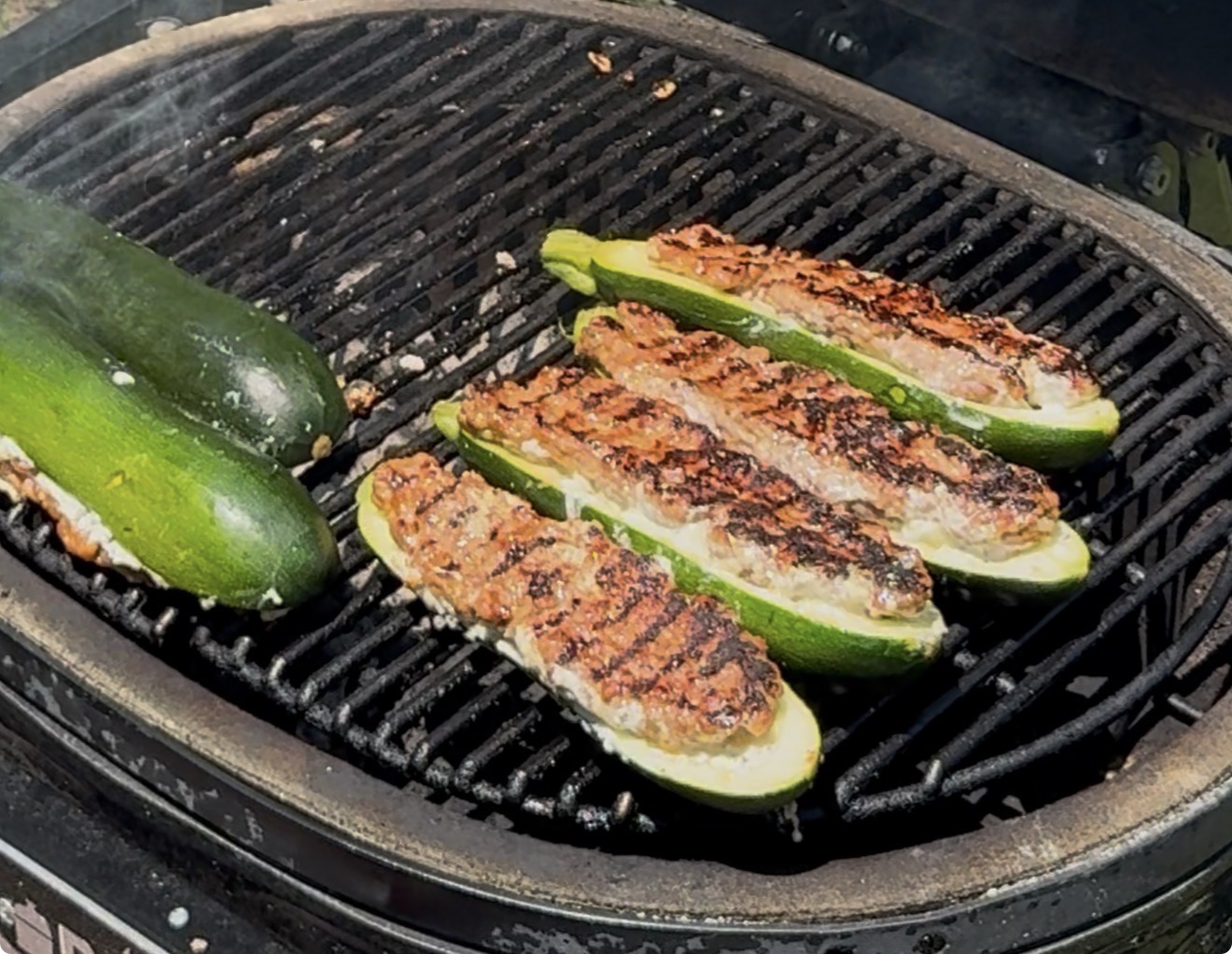 Four zucchini halves stuffed with a cheesy filling and two whole zucchinis are grilling on a barbecue, with grill marks visible and smoke rising.