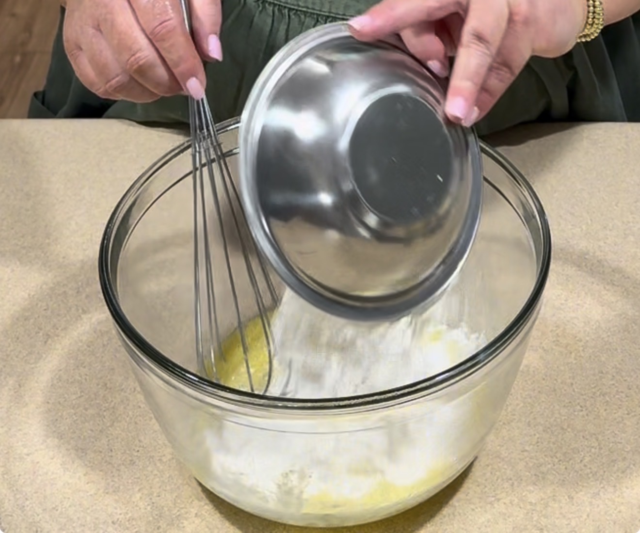 A person pours flour from a metal bowl into a glass mixing bowl with wet ingredients, using a whisk to combine them, on a beige countertop.