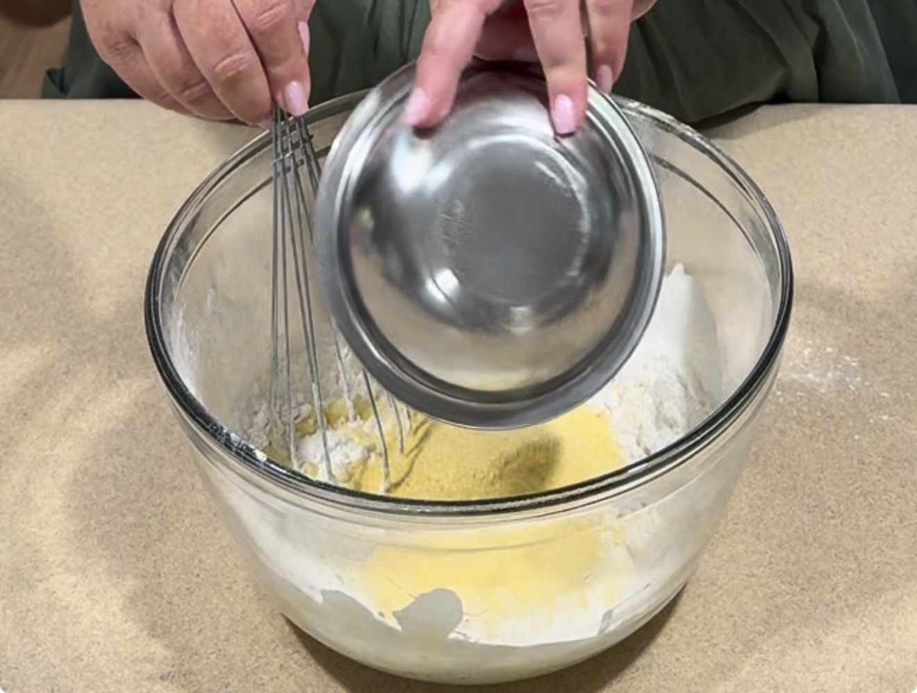A person pours ingredients from a small metal bowl into a glass mixing bowl containing flour and cornmeal, while holding a whisk in the other hand.