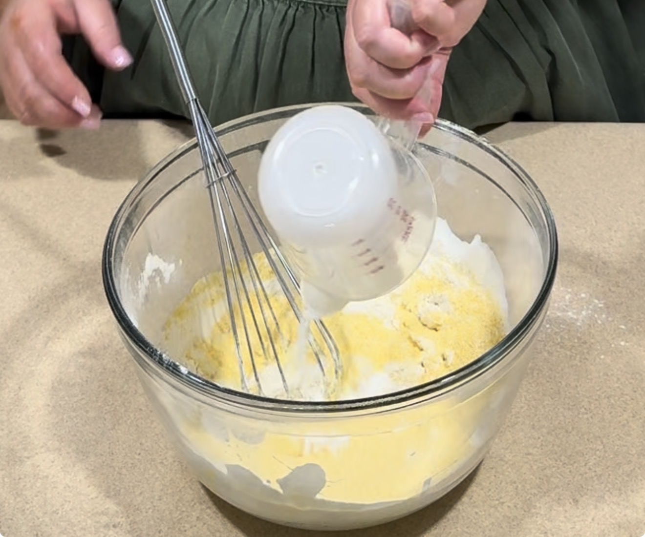 A person pours liquid from a measuring cup into a glass bowl with flour and cornmeal, using a whisk to mix the ingredients on a beige countertop.