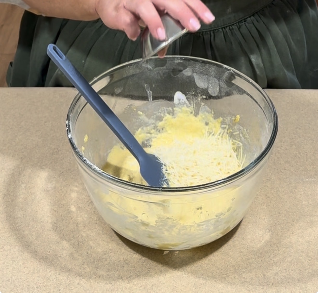 A person pours grated cheese into a glass mixing bowl with yellow batter, using a blue spatula to combine the ingredients on a kitchen countertop.