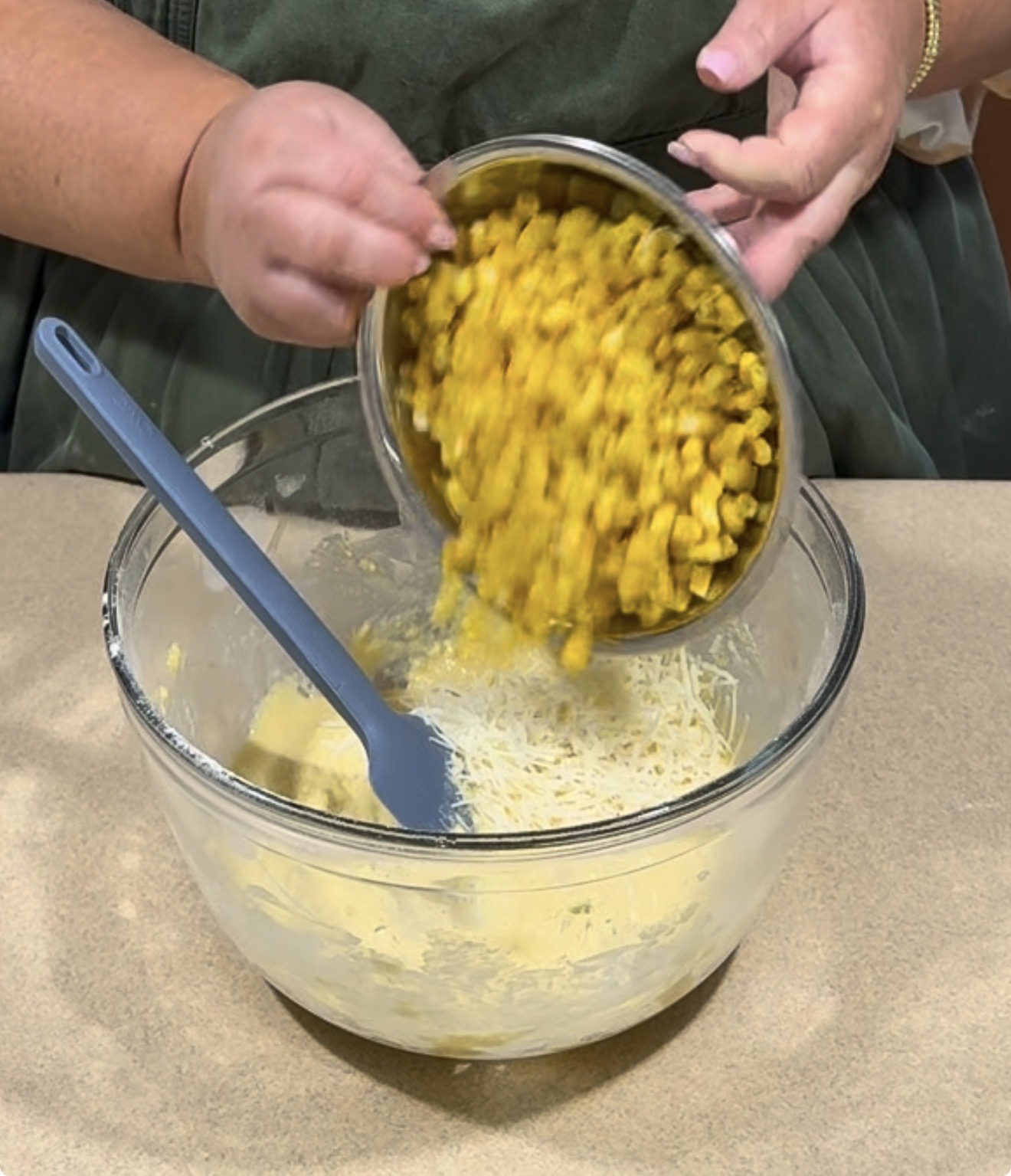 A person pours a bowl of seasoned corn into a glass mixing bowl containing shredded cheese and dough, with a spatula resting inside.