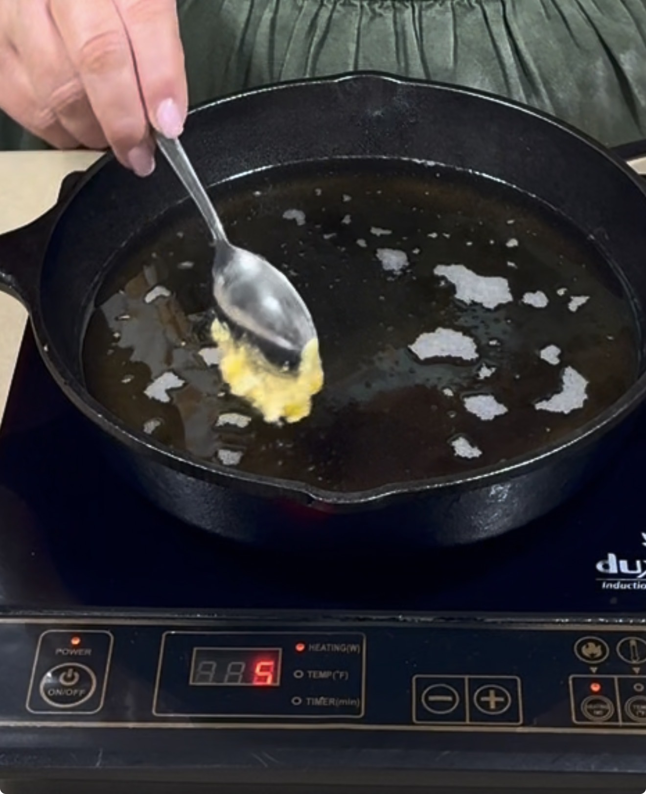 A person holds a spoon with batter above a hot, oiled cast iron skillet on an induction cooktop set to heat level 5.