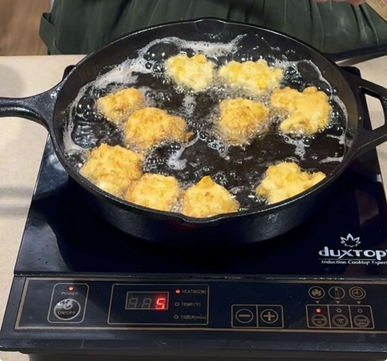 Golden fritters are frying in hot oil in a black cast iron skillet on a portable induction cooktop, with a digital display showing the heat setting at 5.
