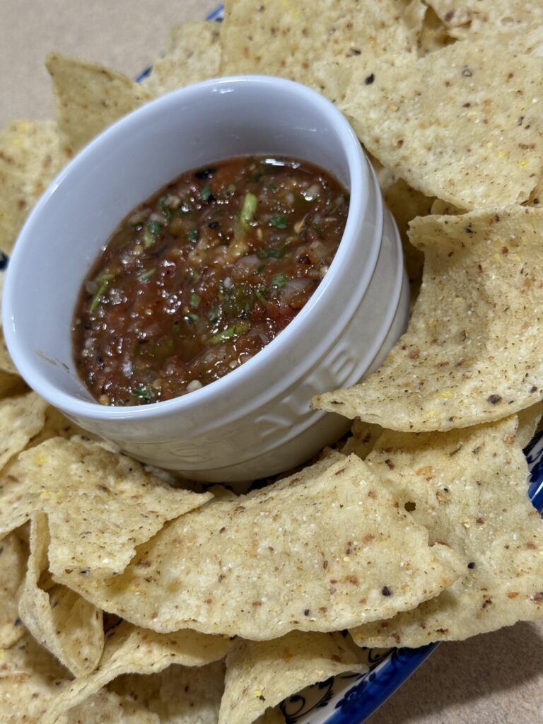 A white bowl filled with chunky salsa is surrounded by crispy tortilla chips on a blue-patterned plate.