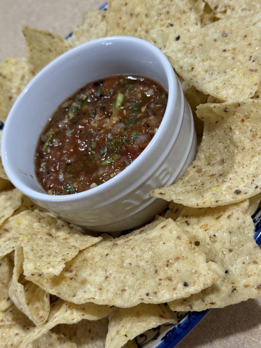 A white bowl filled with chunky salsa is surrounded by crispy tortilla chips on a blue-patterned plate.