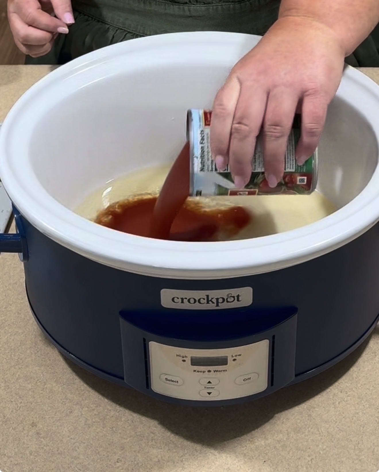 A person pours tomato sauce from a can into a blue Crockpot slow cooker on a kitchen counter.
