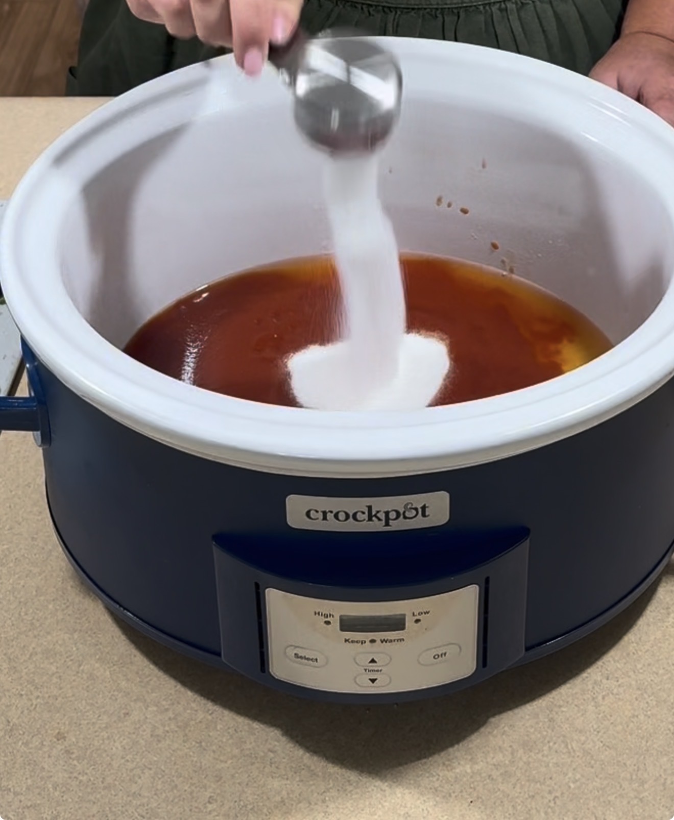 A hand pours white granulated sugar from a metal measuring cup into a blue Crockpot filled with reddish liquid, possibly sauce, on a beige countertop.