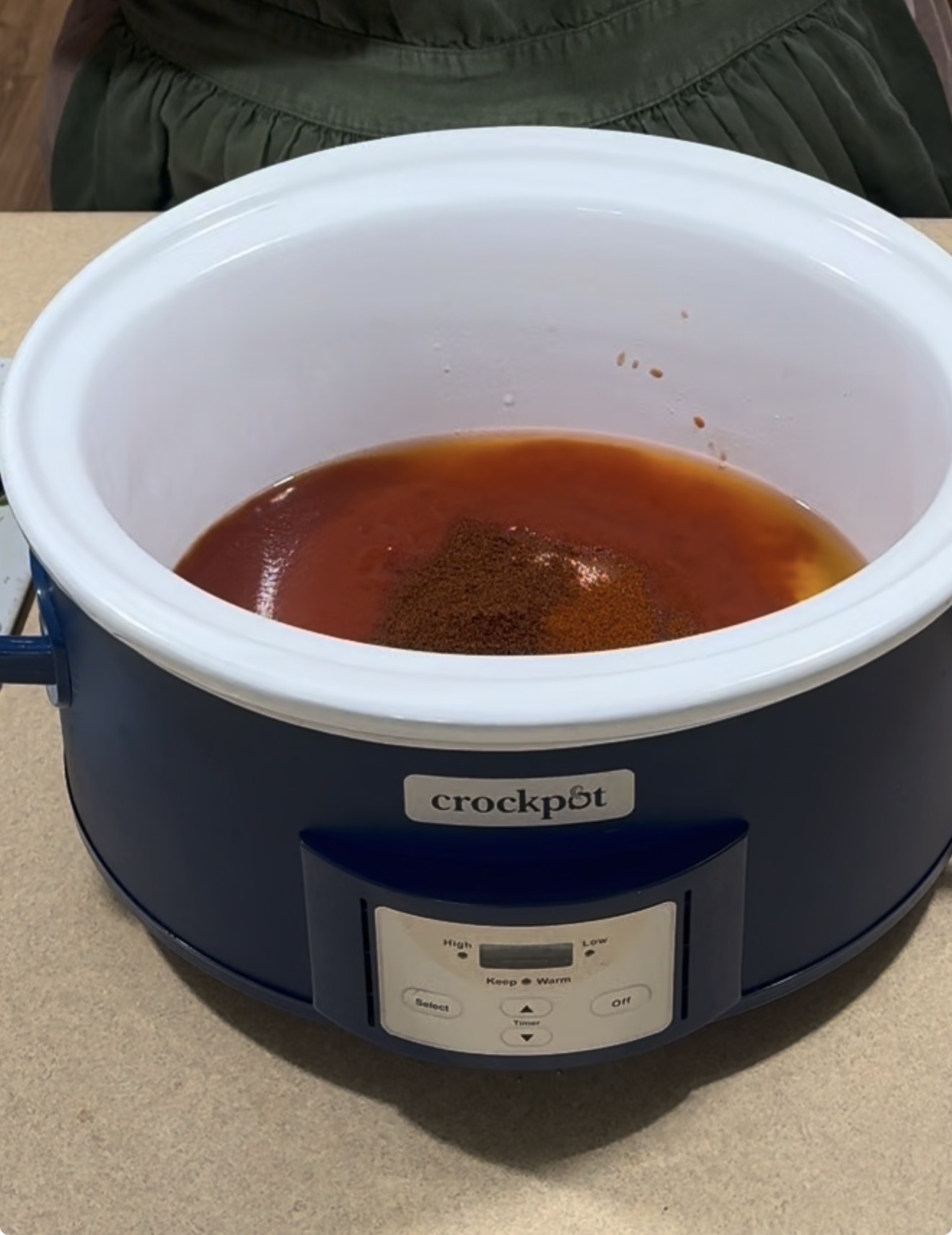 A blue Crock-Pot slow cooker filled with red sauce and seasoning sits on a countertop. A person wearing a green apron stands behind it.