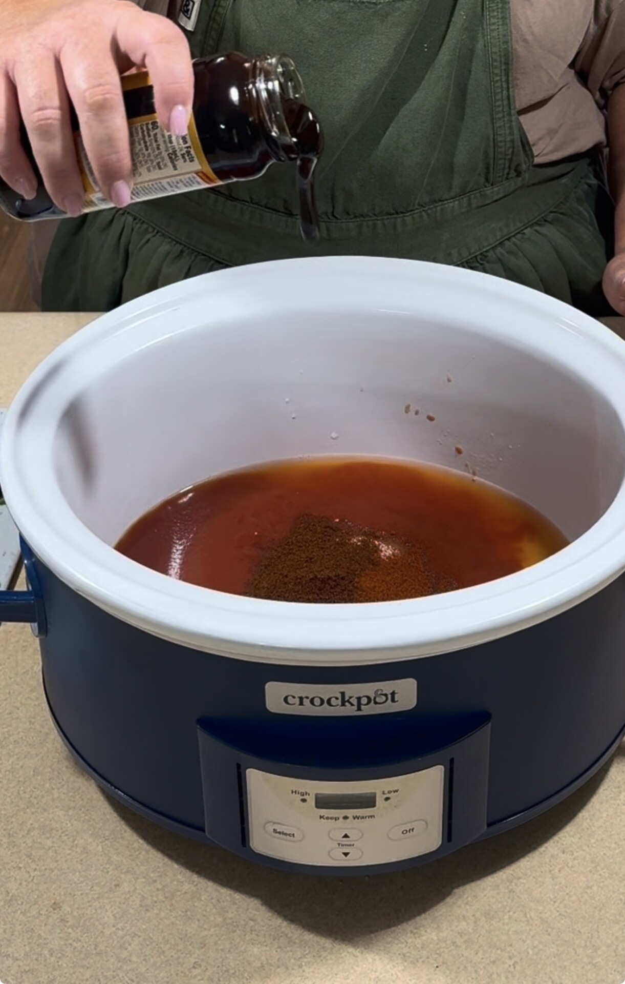 A person pours liquid from a bottle into a blue Crockpot filled with reddish-brown sauce and spices.