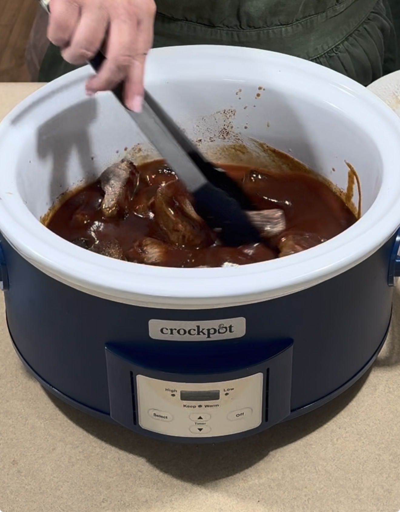 A person uses tongs to stir meat in barbecue sauce inside a blue Crockpot slow cooker on a countertop.