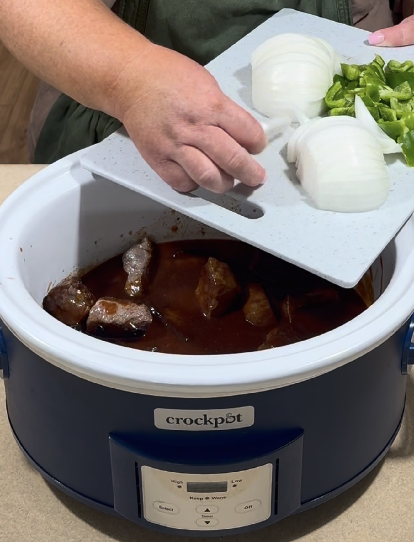 A person adds sliced onions and green peppers from a cutting board into a blue Crockpot filled with chunks of meat in sauce.