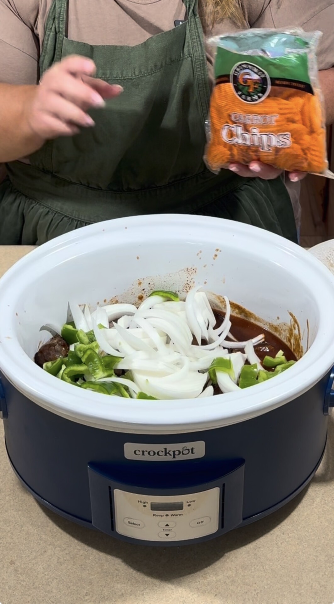 A person in a green apron adds sliced onions and green peppers to a crockpot with sauce, holding a bag of carrot chips next to the slow cooker.