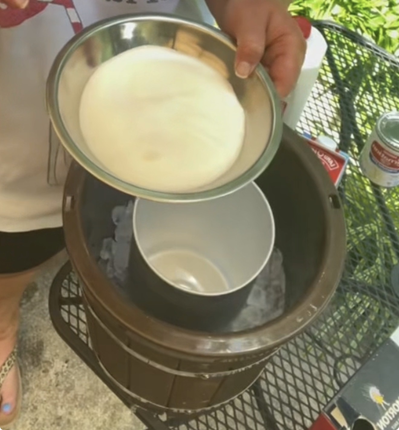 A person holds a metal bowl of cream above an ice cream maker filled with ice, ready to pour the mixture inside. Cans and a box sit on a black outdoor table nearby.