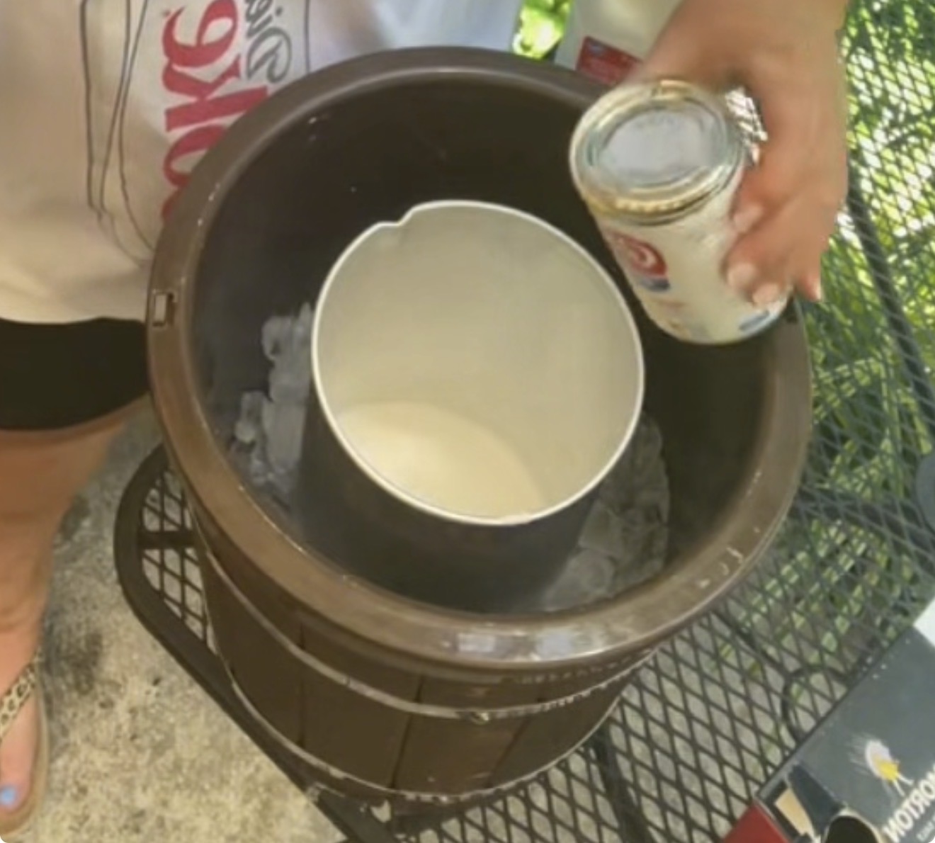 A person pours sweetened condensed milk into an old-fashioned ice cream maker filled with ice, preparing homemade ice cream outdoors.