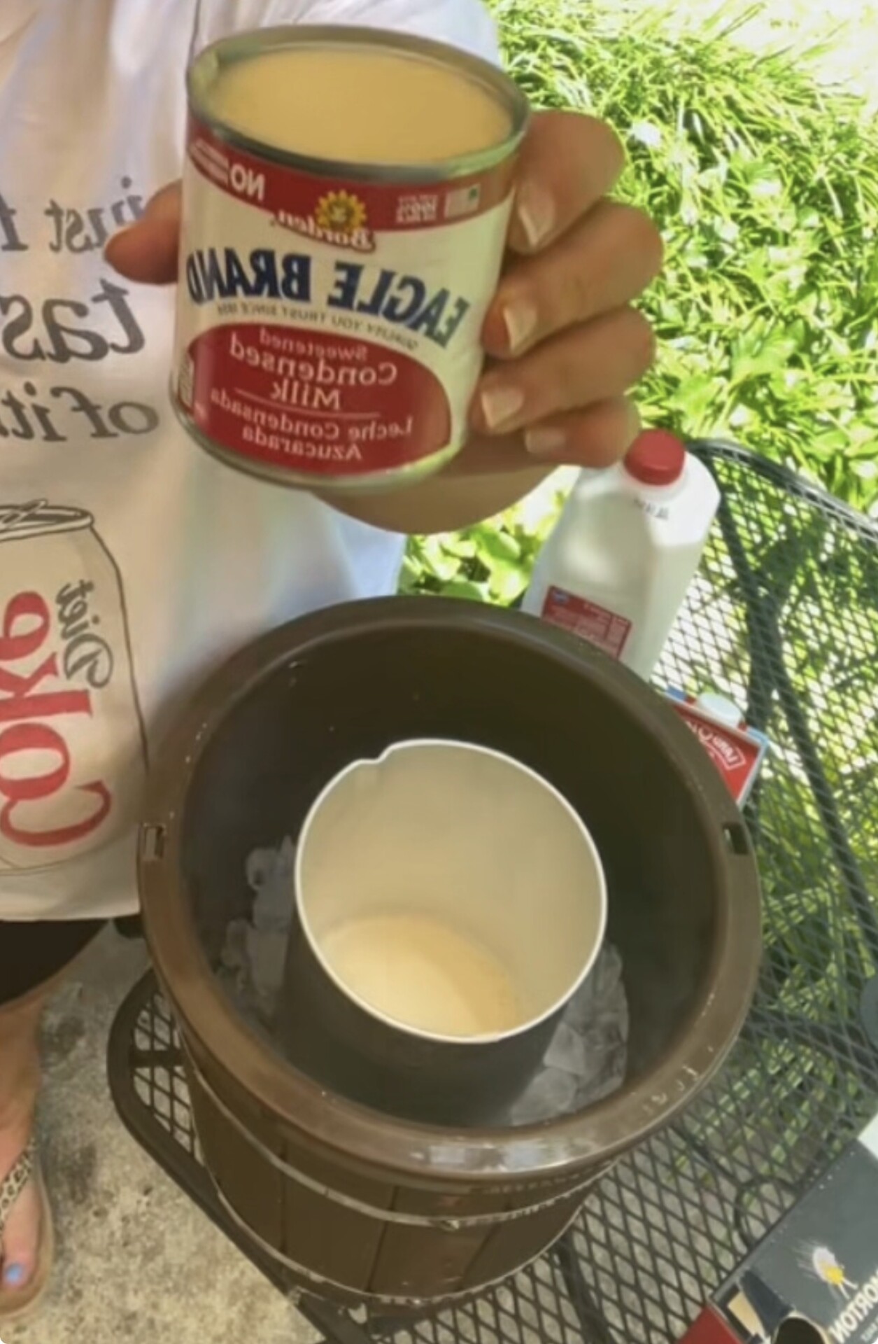 Person holding a can of Eagle Brand sweetened condensed milk above a small ice cream maker with ice and a mixing container inside. A milk jug is on the table outdoors.