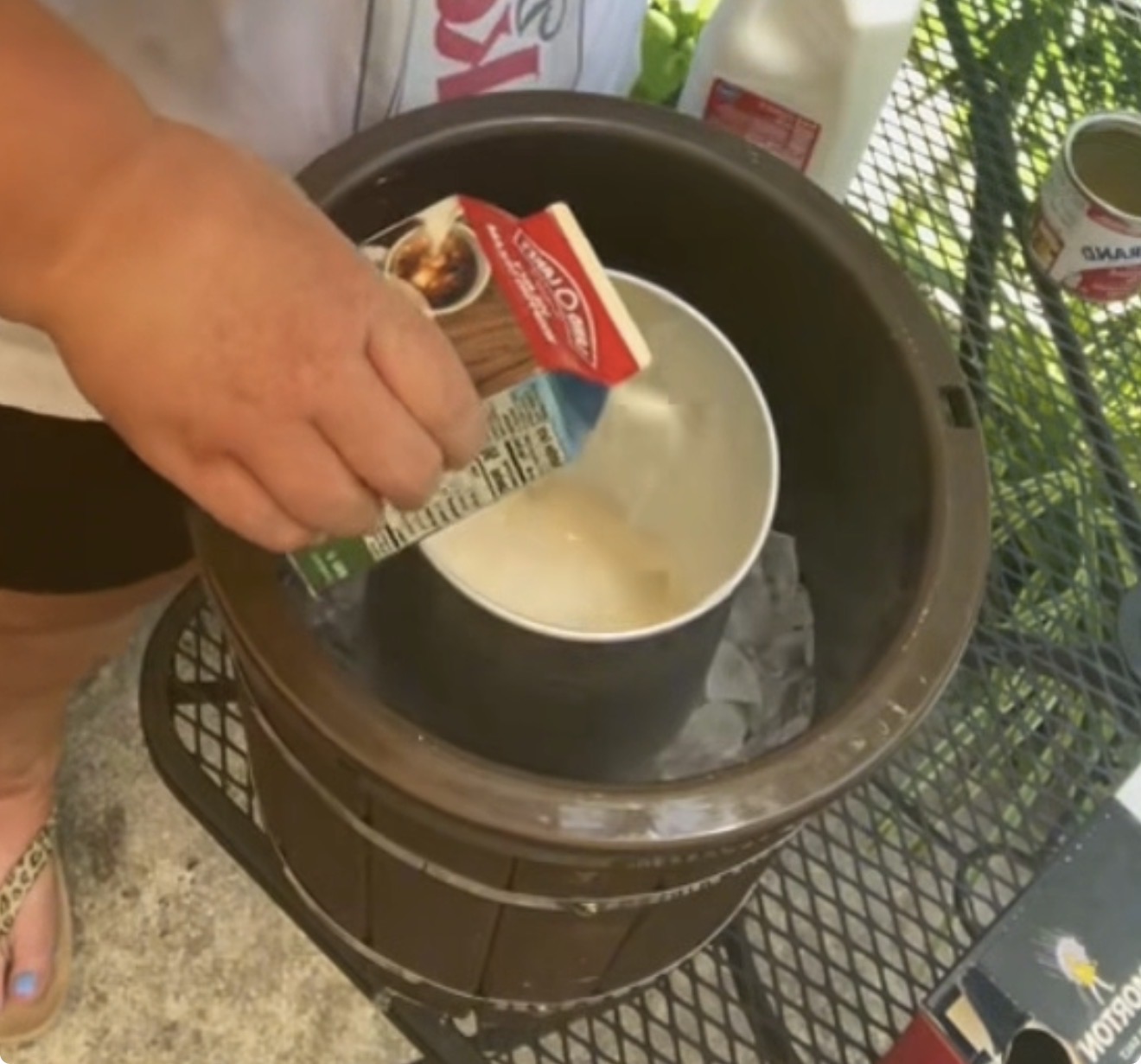 A person pours heavy whipping cream into a metal canister inside an old-fashioned ice cream maker surrounded by ice. Milk and other ingredients are nearby on a metal table.