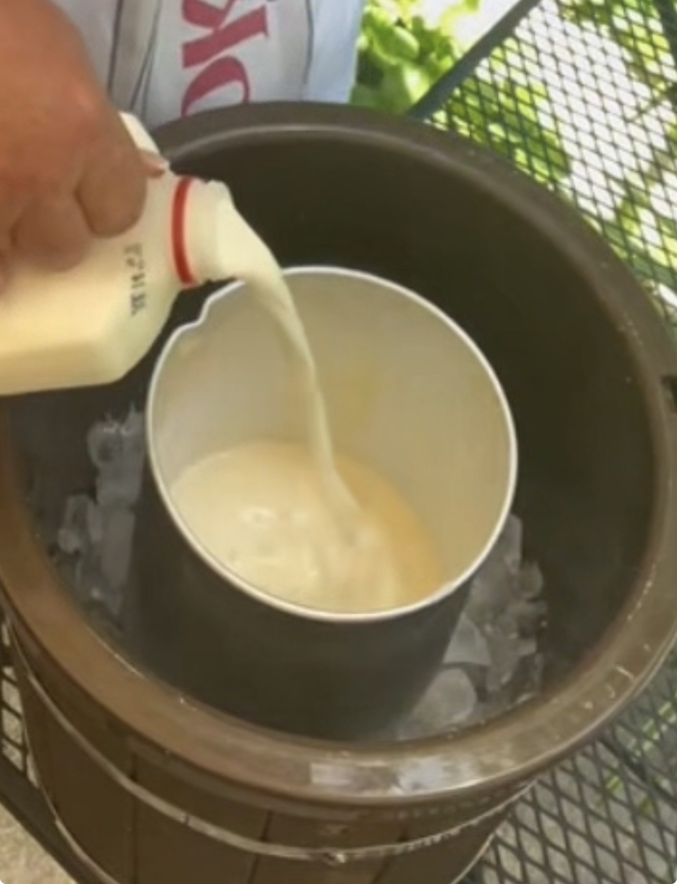 A hand pours milk from a bottle into a metal canister, which sits in a bucket surrounded by ice, possibly preparing homemade ice cream outdoors.