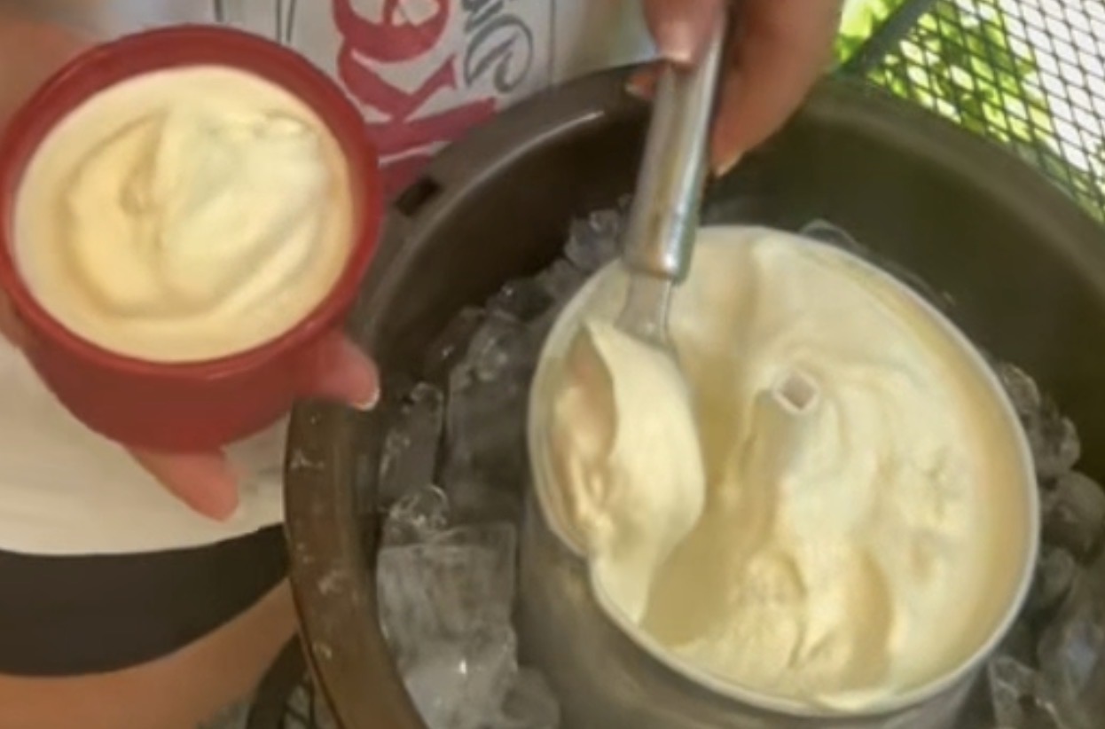 A person scoops homemade vanilla ice cream from an old-fashioned ice cream maker into a small red bowl. The ice cream maker sits in a bucket filled with ice.