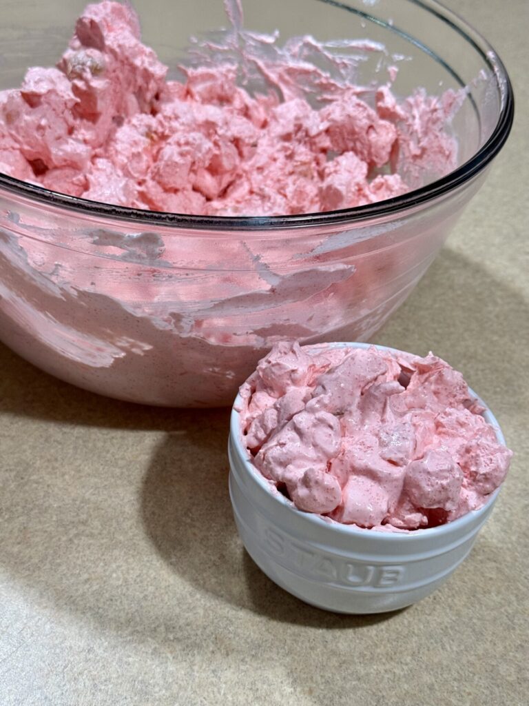 A glass bowl and a small white bowl filled with fluffy pink dessert, possibly strawberry mousse or whipped salad, sit on a beige countertop.