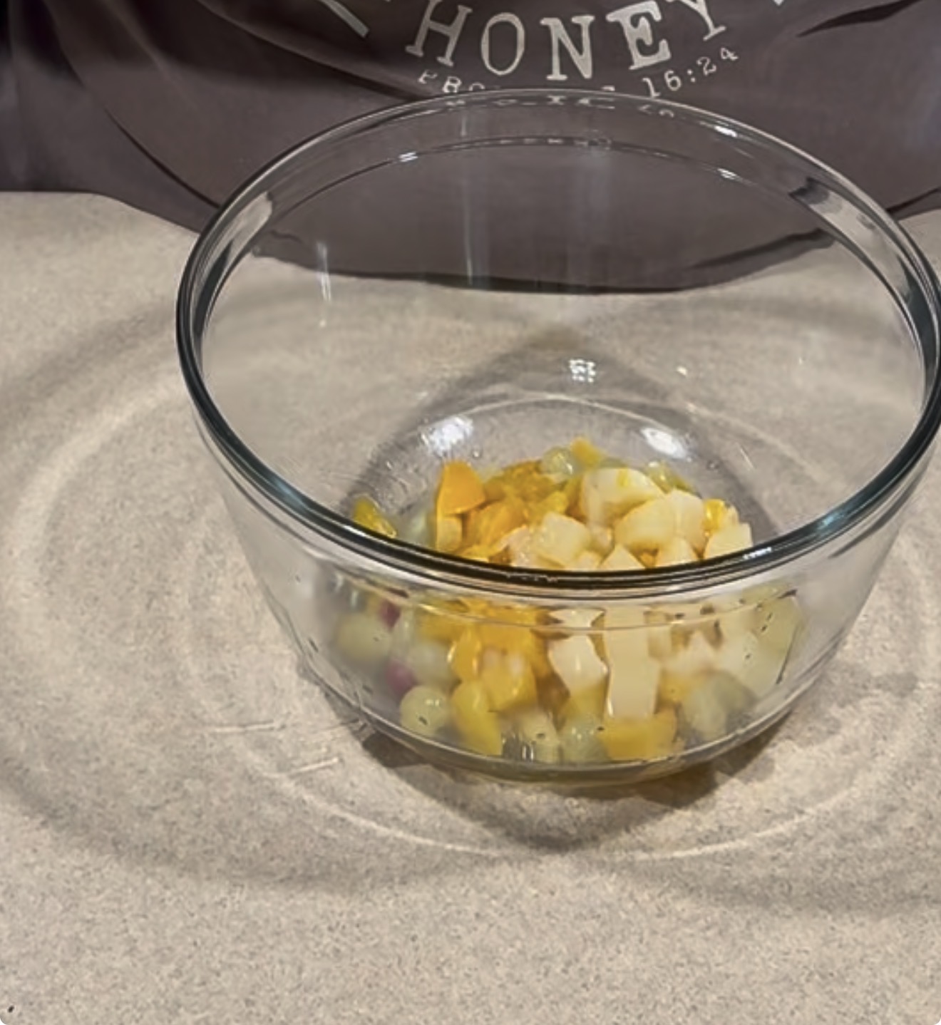 A clear glass bowl filled with chopped fruit pieces, including yellow and white cubes, sits on a light countertop. A gray cloth is in the background.