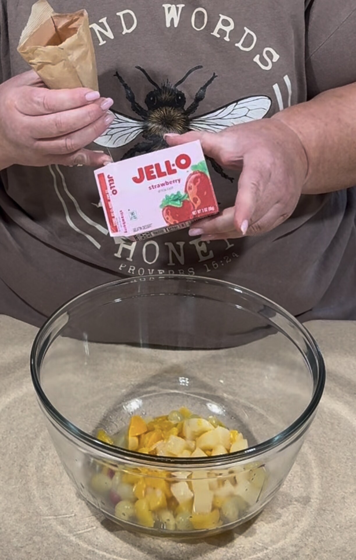 A person holding strawberry Jello mix and a paper pouch above a glass bowl containing mixed canned fruit. The person wears a brown shirt with a bee illustration and the words KIND WORDS HONEY.
