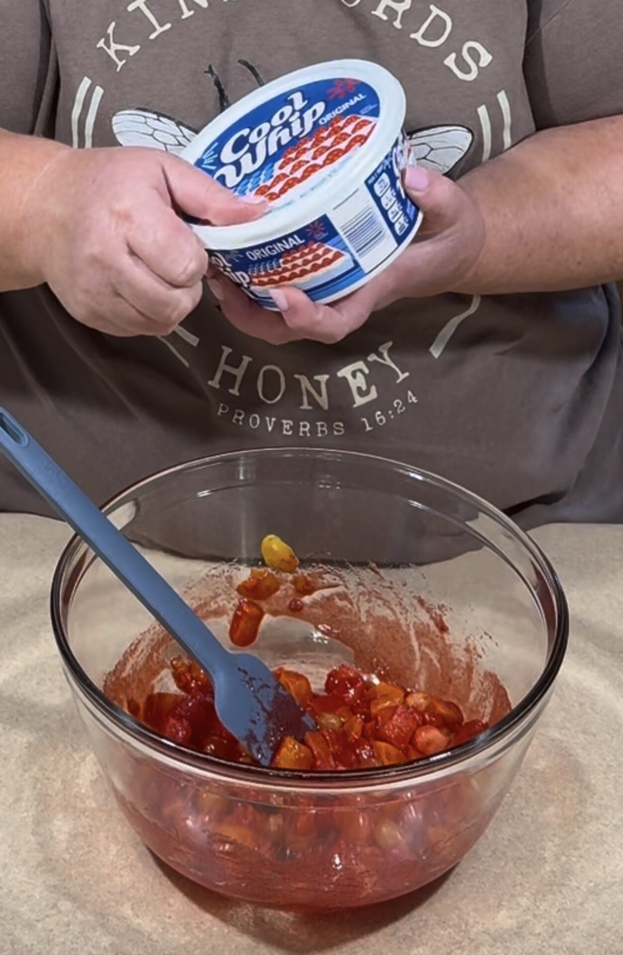 Person holding a container of Cool Whip above a glass bowl of fruit salad with a spatula inside. The individual is wearing a T-shirt with text and a butterfly graphic.