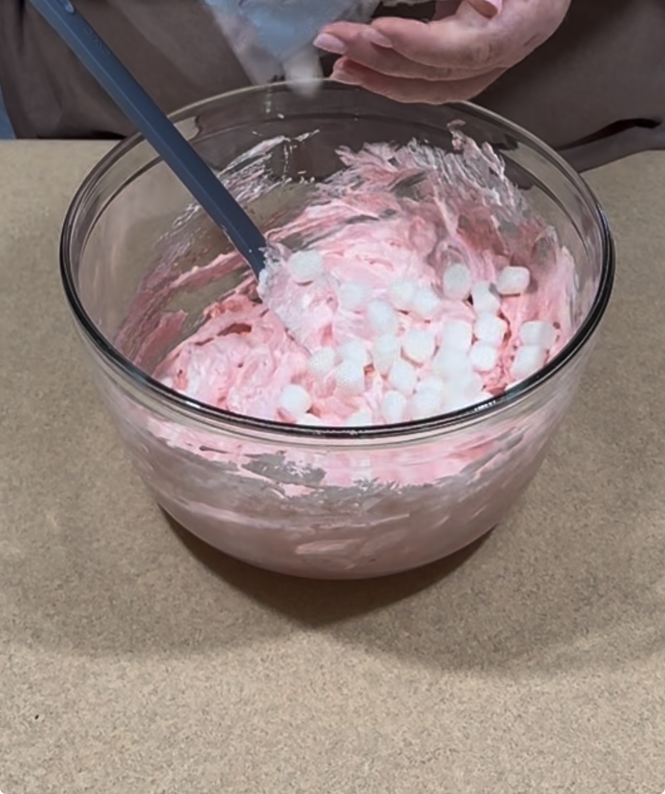A person mixes small white marshmallows into a large glass bowl of pink batter using a blue spatula. The bowl sits on a beige countertop.