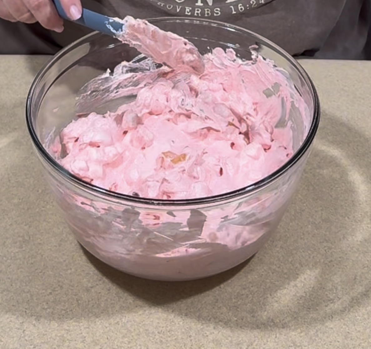 A person is mixing a large glass bowl of fluffy pink whipped mixture with a spatula on a beige countertop.