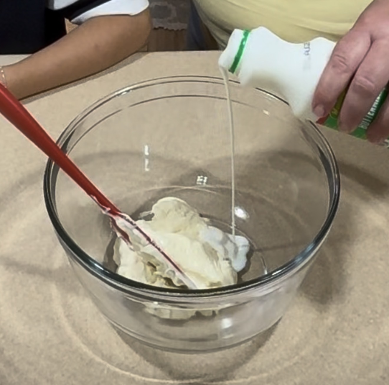 A person pours liquid from a bottle into a glass bowl containing a creamy mixture, using a red spatula. Another person’s arm is visible beside the bowl on a beige surface.