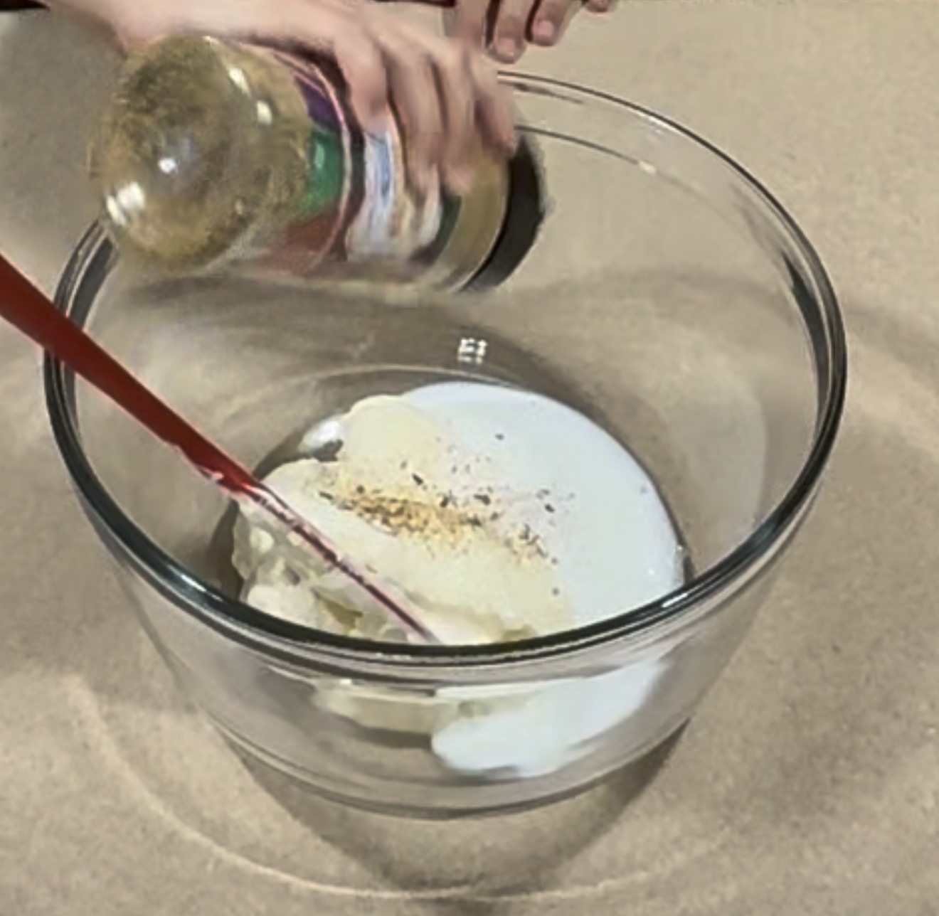 A hand sprinkles seasoning from a bottle into a clear bowl containing mayonnaise and milk, with a red spatula resting inside.