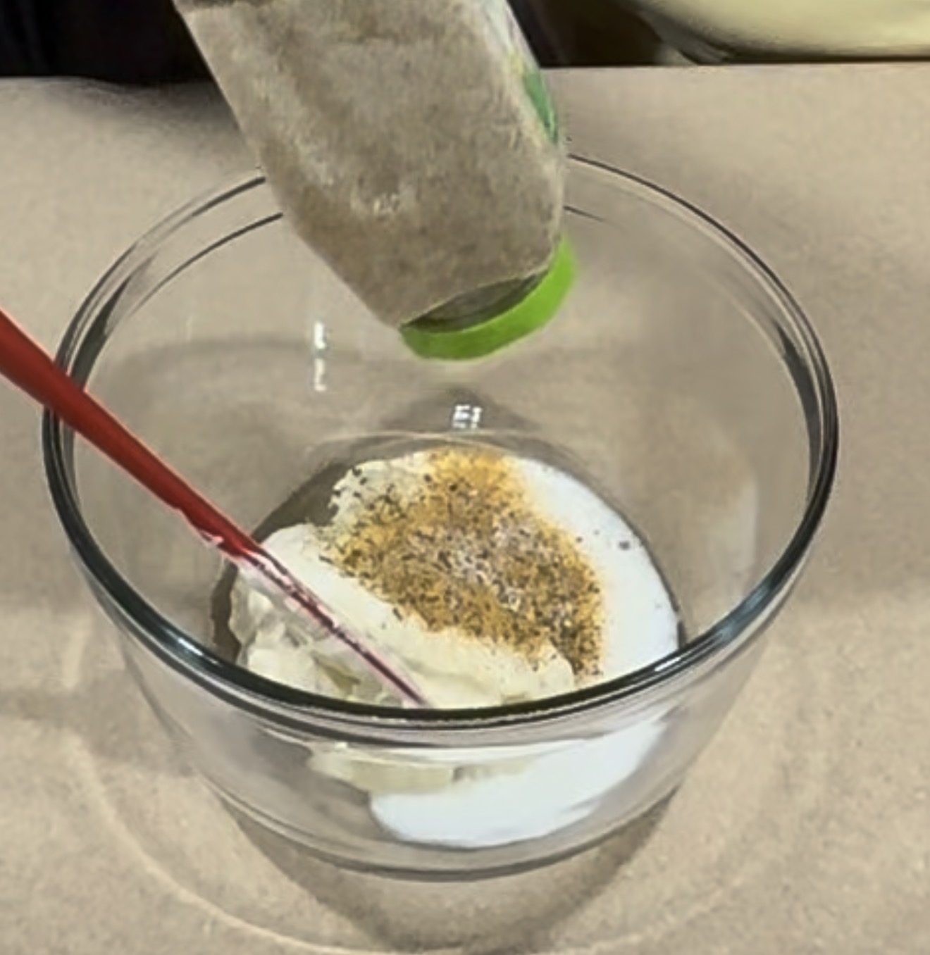 A spice jar with a green lid is held upside down over a glass mixing bowl, sprinkling seasoning onto a mixture of white creamy ingredients like mayonnaise and milk, with a red spatula resting inside the bowl.