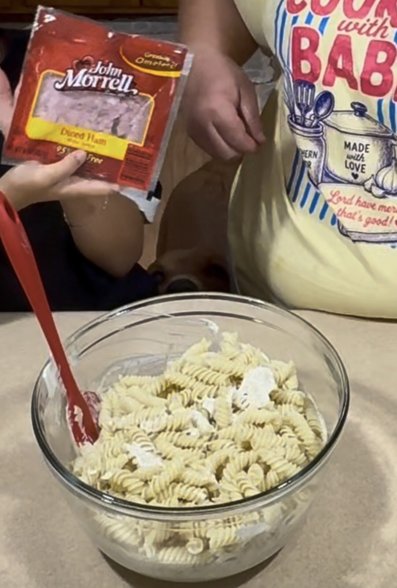 Two people stand near a bowl of rotini pasta being mixed with a red spatula; one person holds a package of John Morrell diced ham while the other wears a shirt with a baking-themed design.