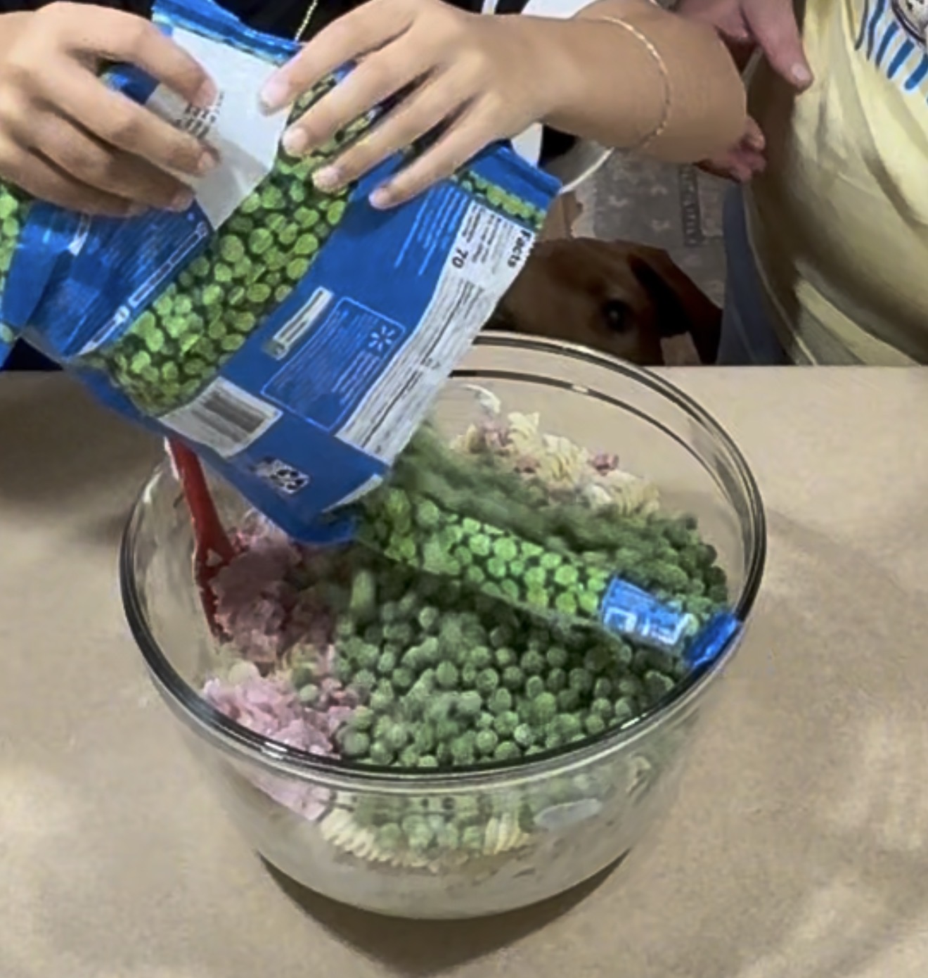 Two people pour frozen peas from a blue bag into a clear bowl filled with pasta and ham, preparing food on a kitchen counter. A dog looks up from the floor in the background.