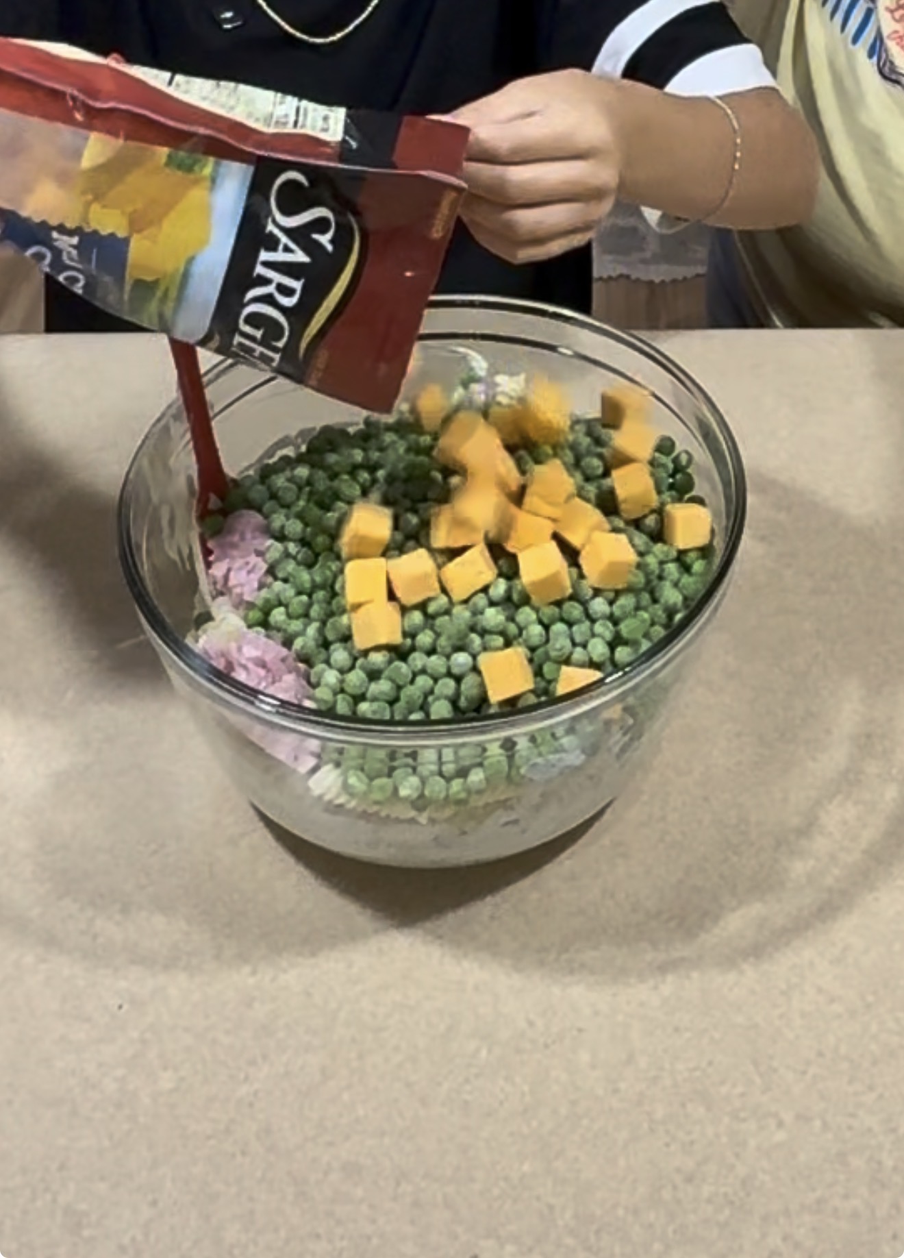 Two people prepare food by adding cubes of cheddar cheese from a Sargento bag into a glass bowl filled with frozen peas and other ingredients on a counter.