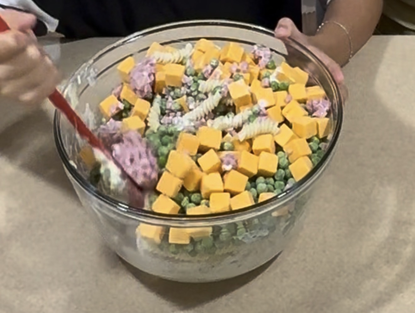 A person mixes a large glass bowl filled with rotini pasta, cubed cheese, green peas, and some pink and white ingredients using a red spatula.