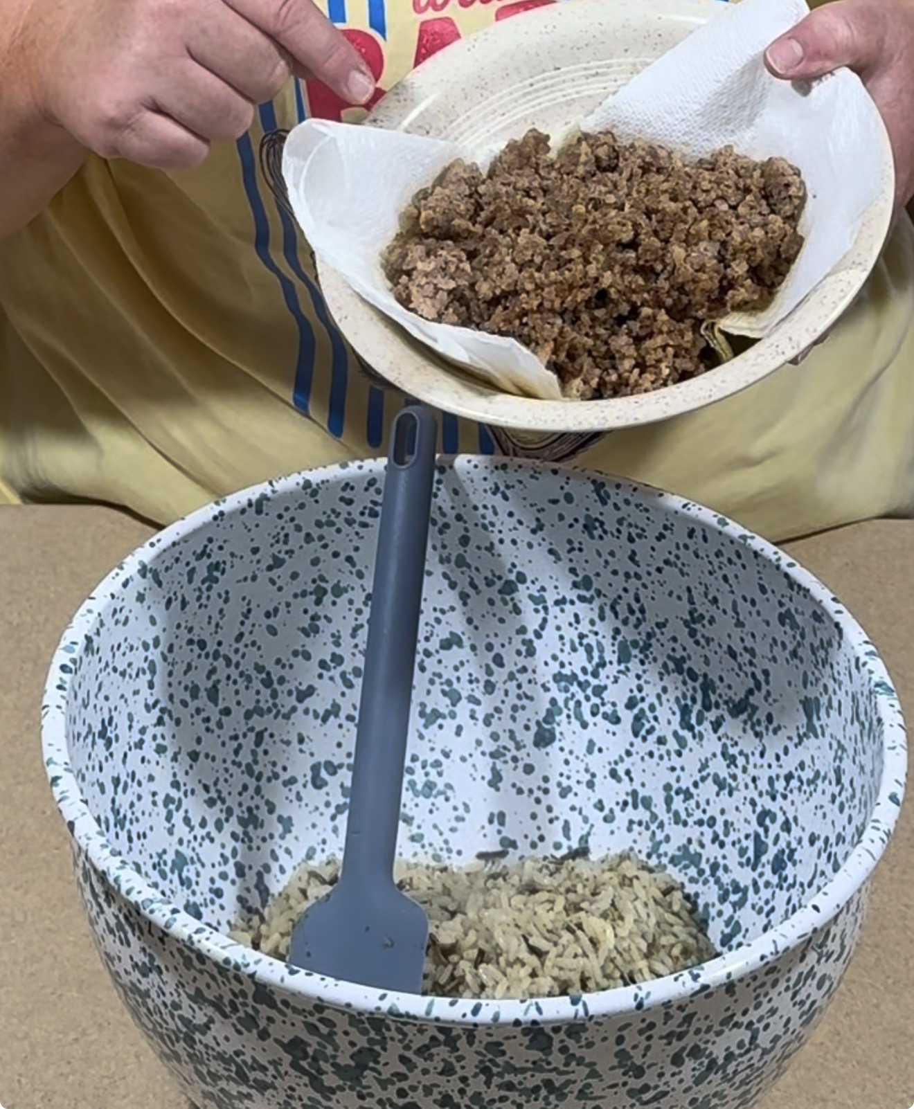 A person pours cooked ground beef from a bowl lined with paper towels into a large speckled mixing bowl containing rice, using a spatula.