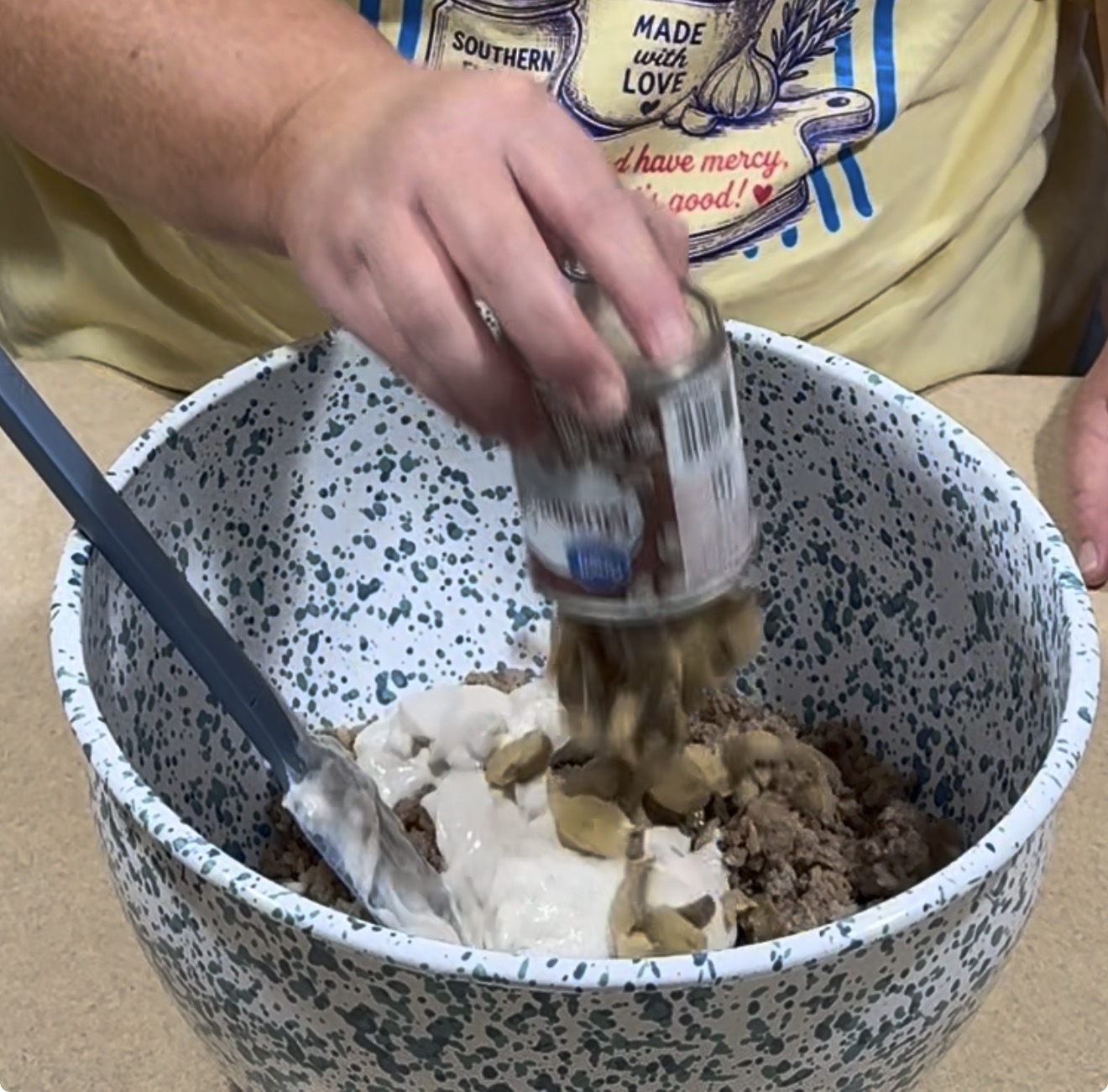 A hand pours canned mushrooms into a speckled mixing bowl with cream soup and ground meat. A spatula is in the bowl. The person wears a yellow shirt with food-related graphics and text.