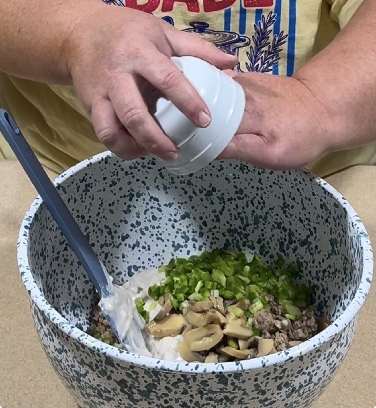 A person adds chopped ingredients from a small white bowl into a large speckled mixing bowl filled with ground meat, mushrooms, chopped green peppers, and a creamy mixture. A spatula rests inside the bowl.