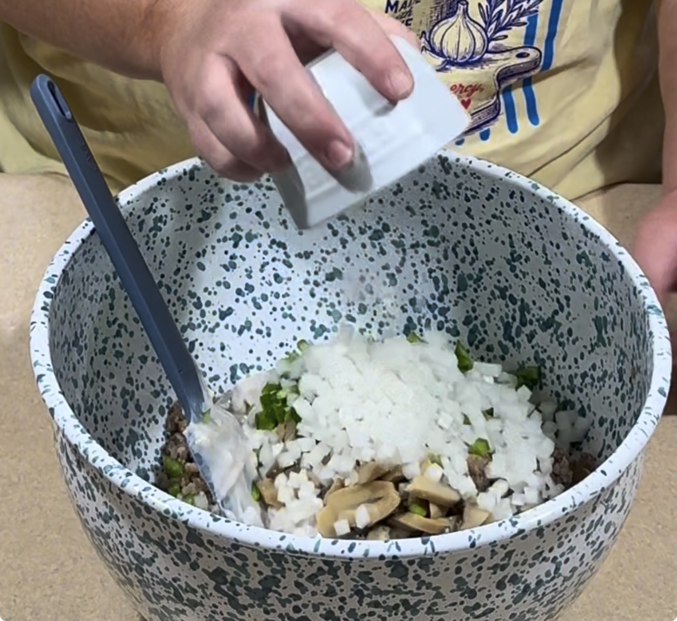 A person pours chopped onions from a small bowl into a speckled mixing bowl containing mushrooms, ground meat, and green peppers. A blue spatula rests in the bowl.