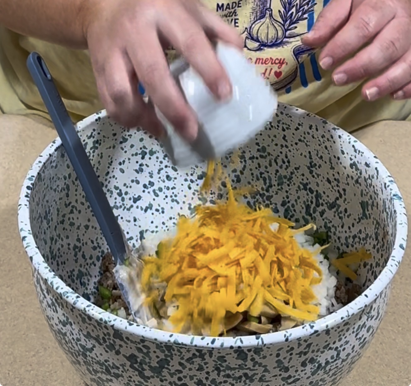 A person pours shredded cheddar cheese from a white bowl into a large speckled mixing bowl containing various ingredients, including ground meat and sour cream. A blue spoon rests in the mixing bowl.