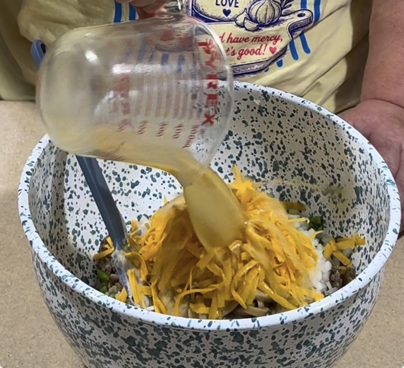 A person pours liquid from a measuring cup into a speckled bowl filled with shredded cheese, onions, and other ingredients, while mixing with a blue spatula.
