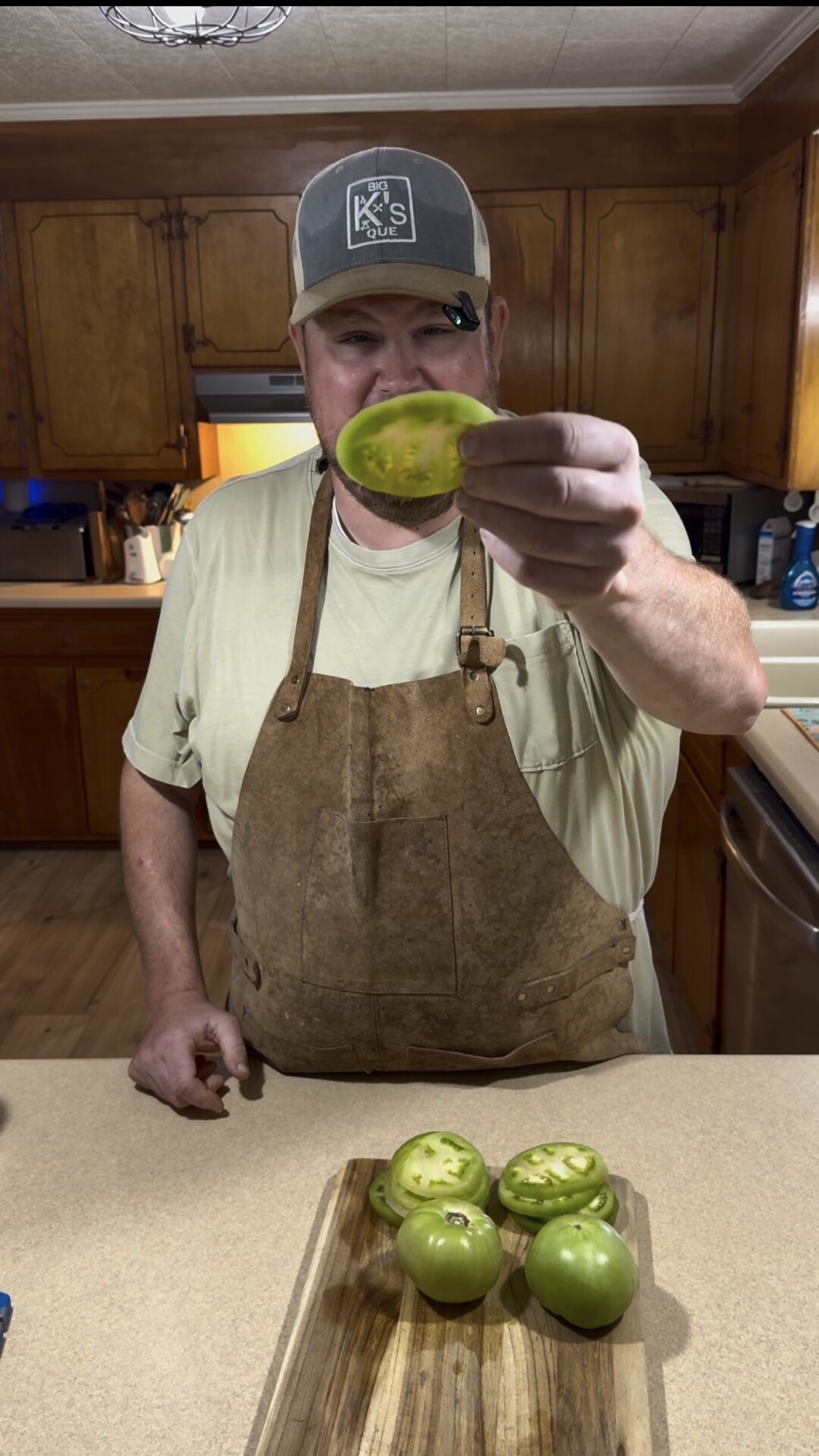 A person wearing a brown apron and a cap stands in a kitchen, holding up a slice of green tomato. Other green tomato slices are on a wooden cutting board on the counter.
