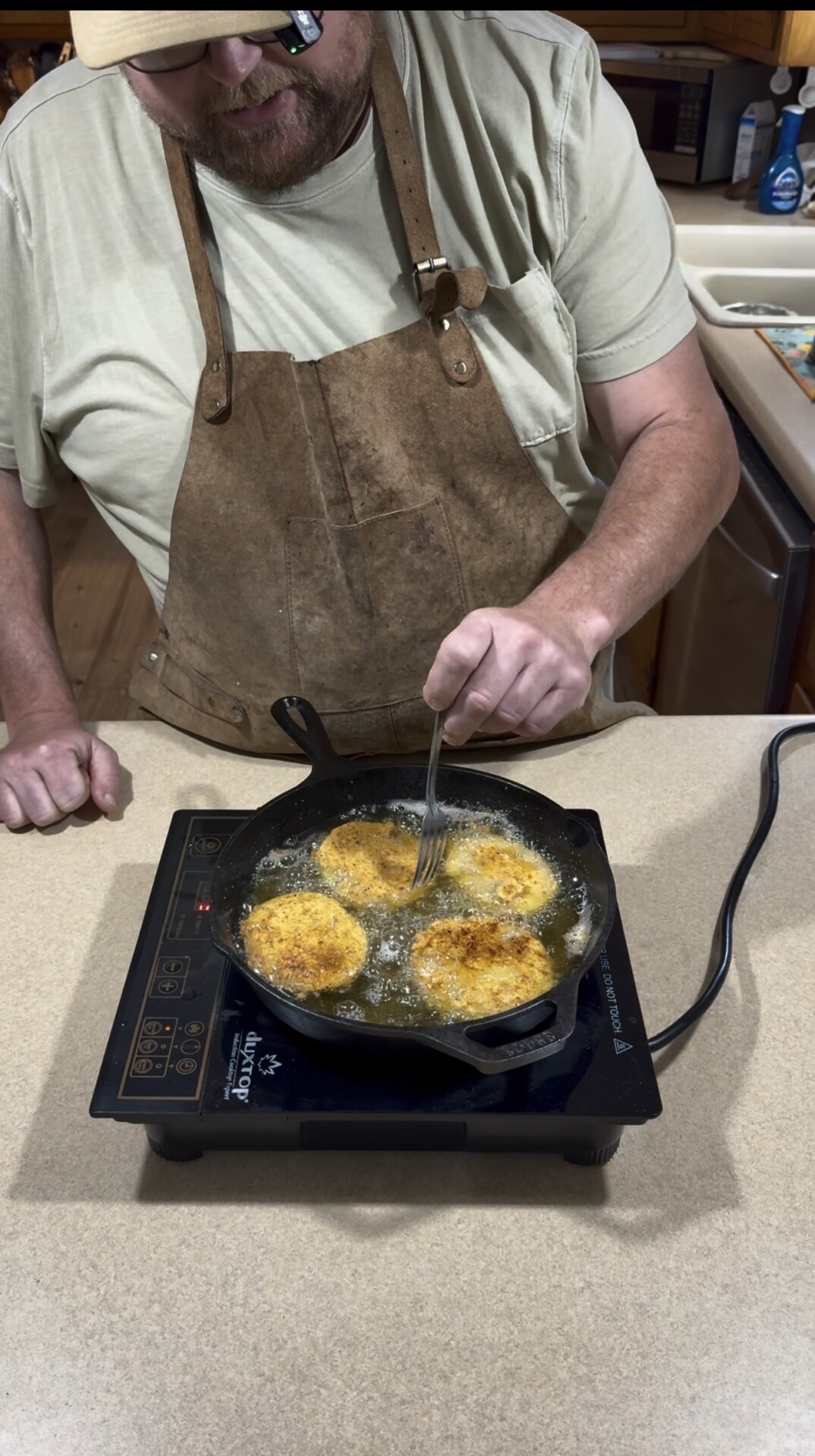 A person in a tan shirt and brown apron is frying four breaded slices in a cast-iron skillet on an electric cooktop, using a fork to turn the food. The scene is set in a kitchen.