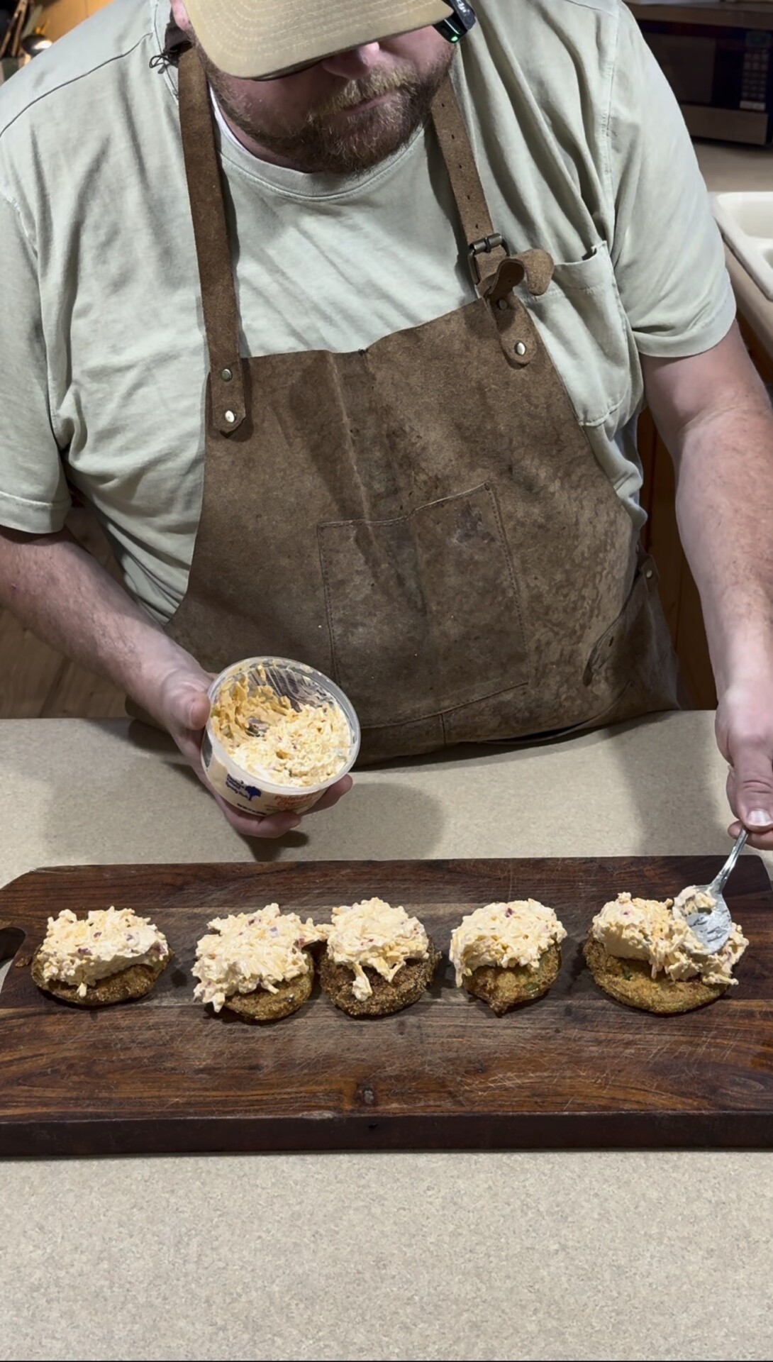 A person wearing a brown apron is spooning a creamy spread onto five fried green tomato slices arranged in a row on a wooden board.