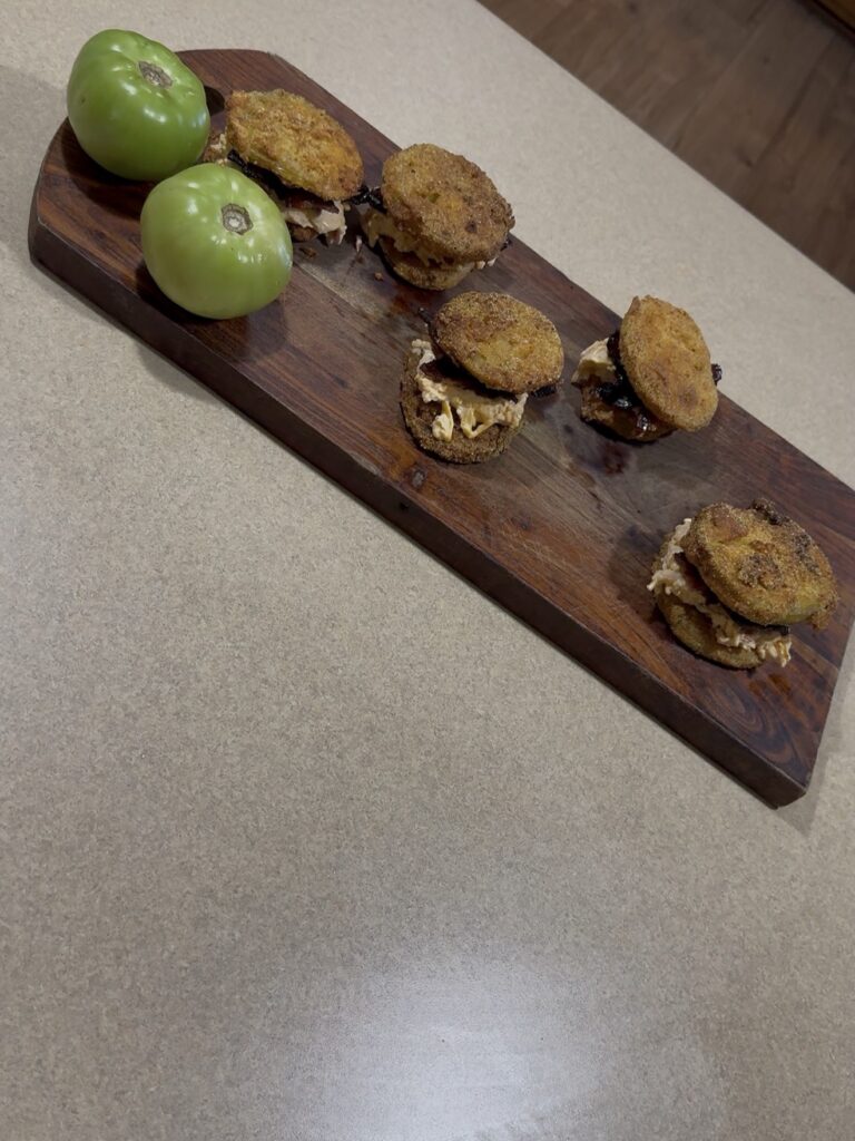 A wooden serving board with five fried green tomato sandwiches and two whole green tomatoes, placed on a beige countertop.