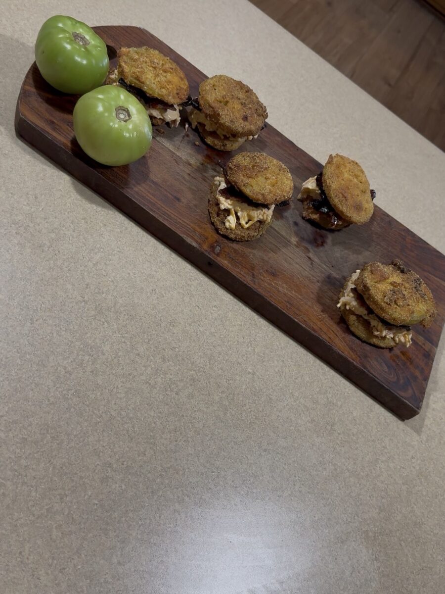 A wooden serving board with five fried green tomato sandwiches and two whole green tomatoes, placed on a beige countertop.