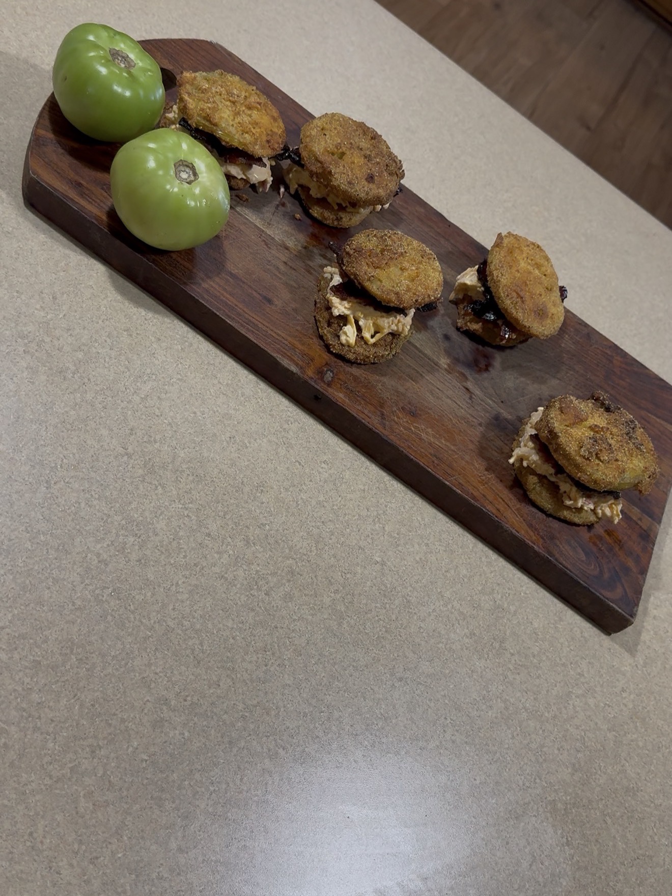 A wooden serving board with five fried green tomato sandwiches and two whole green tomatoes, placed on a beige countertop.