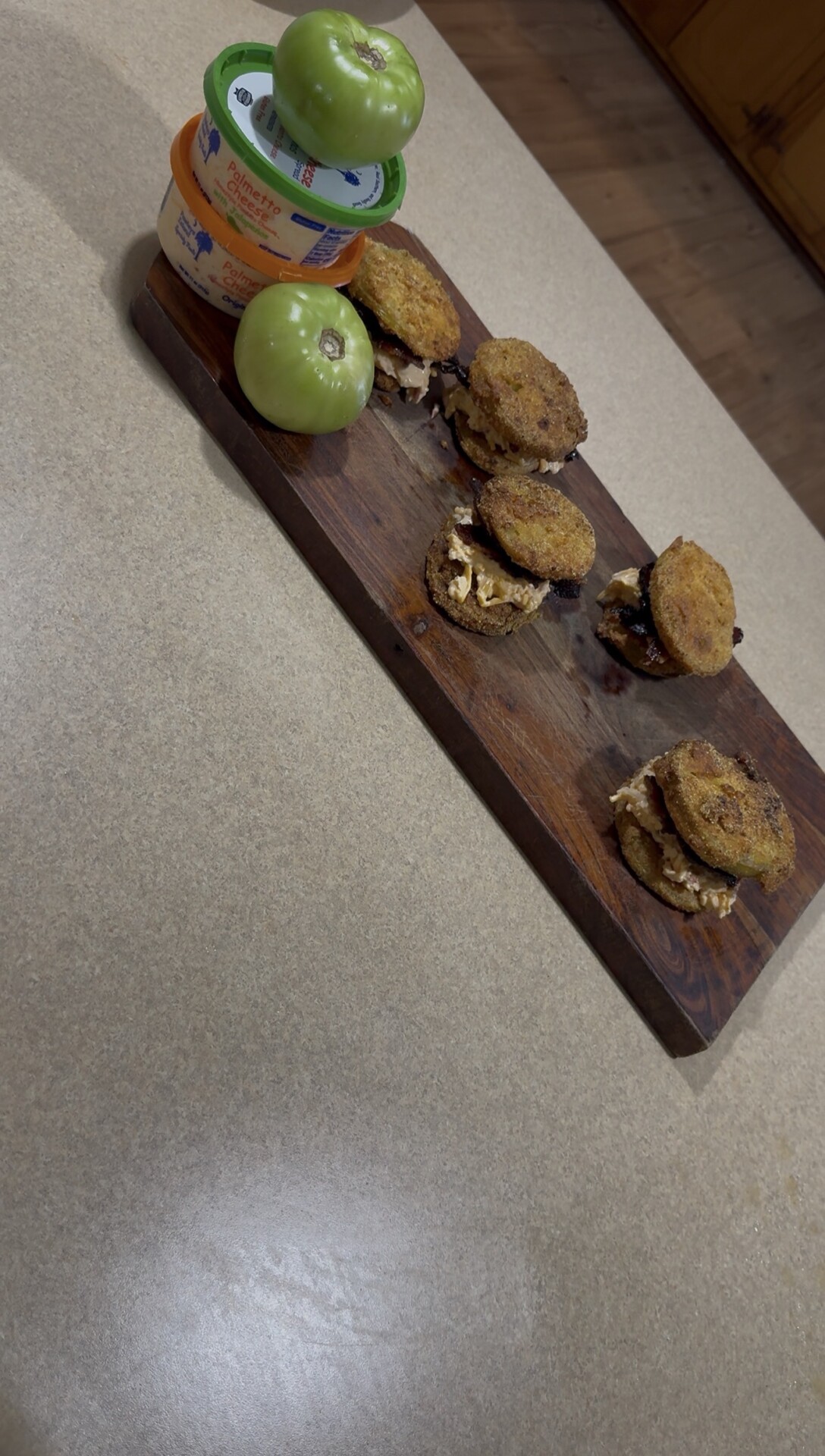 A wooden board with five fried green tomato sandwiches, a whole green tomato, and two containers of pimento cheese is placed on a beige countertop.